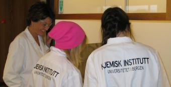 two girls in white laboraory coats are guided in the lab by an engineer, also wearing a lab coat 