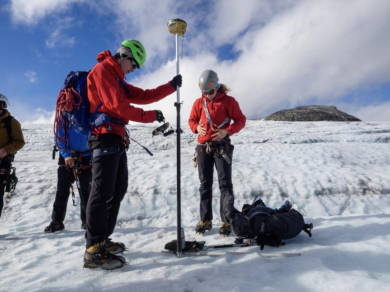 To studenter i front står på Austedalsbreen iført fullt sikkerhetsutstyr som hjelm, sekk med tau i og isspiggsko. Dei samlar inn data ved hjelp av ein drone (UAV)