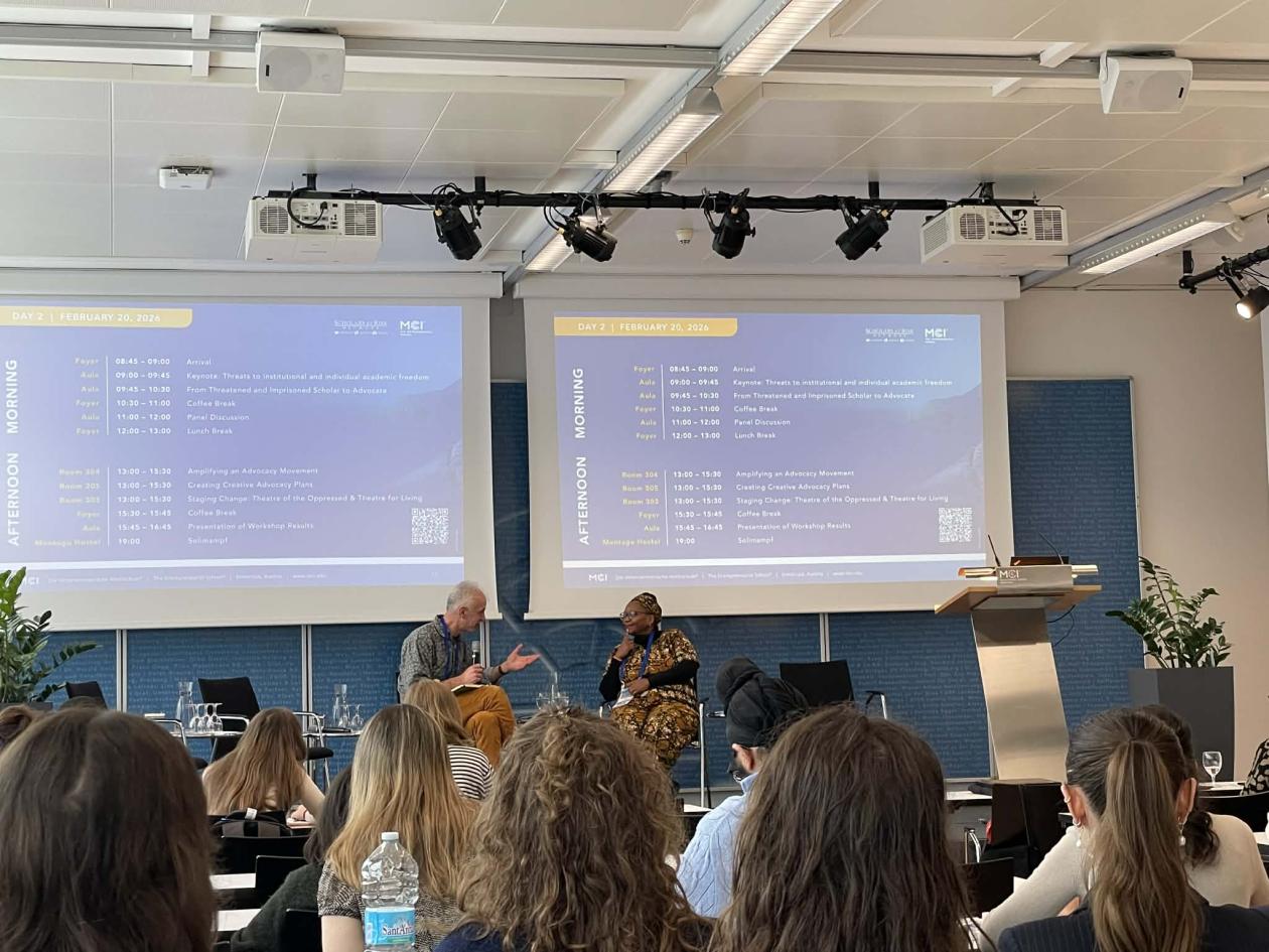 Two people on a stage talking, the screens behind them shows the conference program