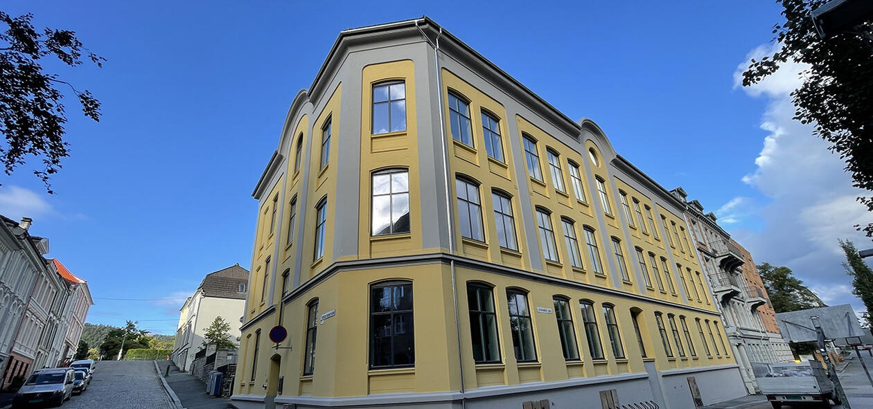 Fasaden til huset Ulrike Pihls hus. Gulmalt med blåhimmel i bakgrunnen. Invitasjon med hvitebokstaver står øverst på den blå himmelen.