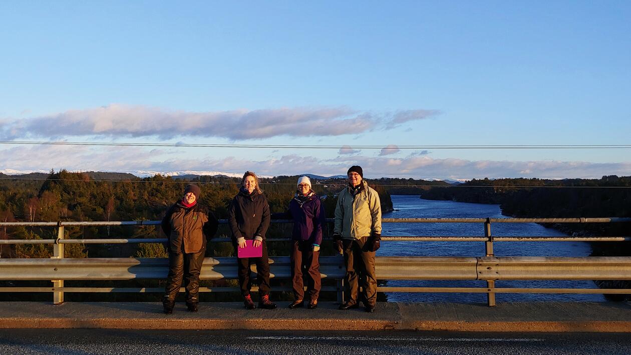 Four of the project members standing on the bridge across the Fonnes stream at sunset, blue skies and the Fonnes stream in the background.
