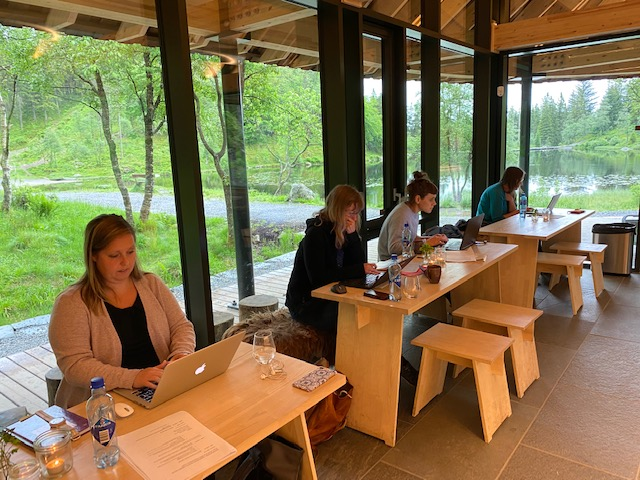 Four women working behind desks with a trees, grass and a lake seen through the window behind them