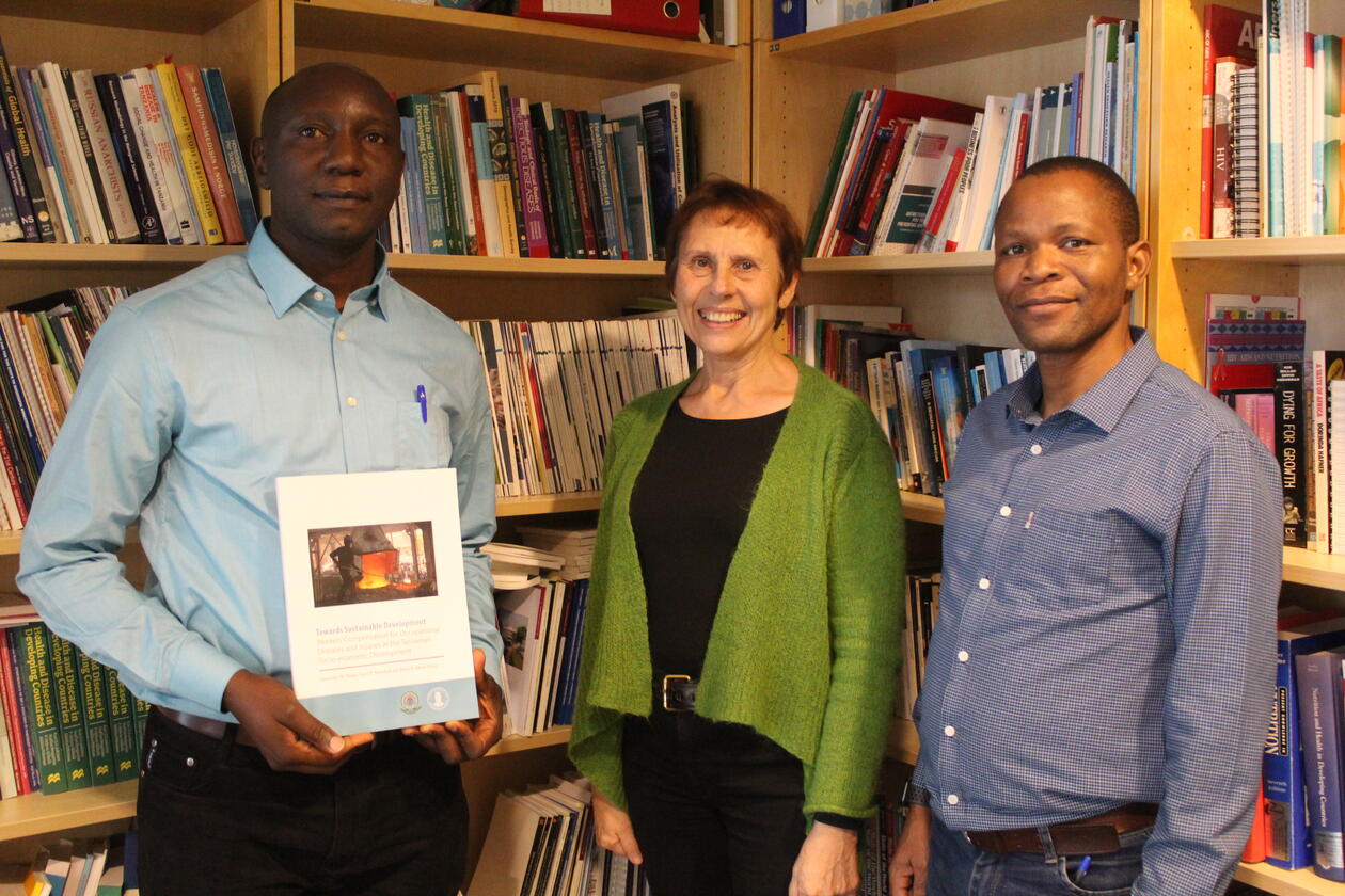 Three people posing in front of a book shelf with books. The person on the left holds a book. 