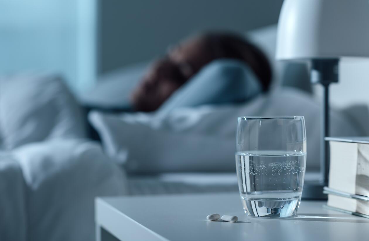 glass med vann og medisiner i forgrunnen - i bakgrunnen, ute av fokus, en kvinne som ligger på en seng og sover eller hviler 