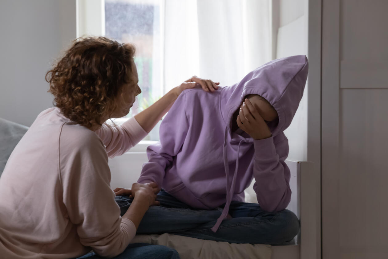 Tenåring med fiolett hettejakke stitter på sengen og blir trøstet av sin mor som holder henne i skulderen og på armen. Tenåringen har hånden foran ansiktet