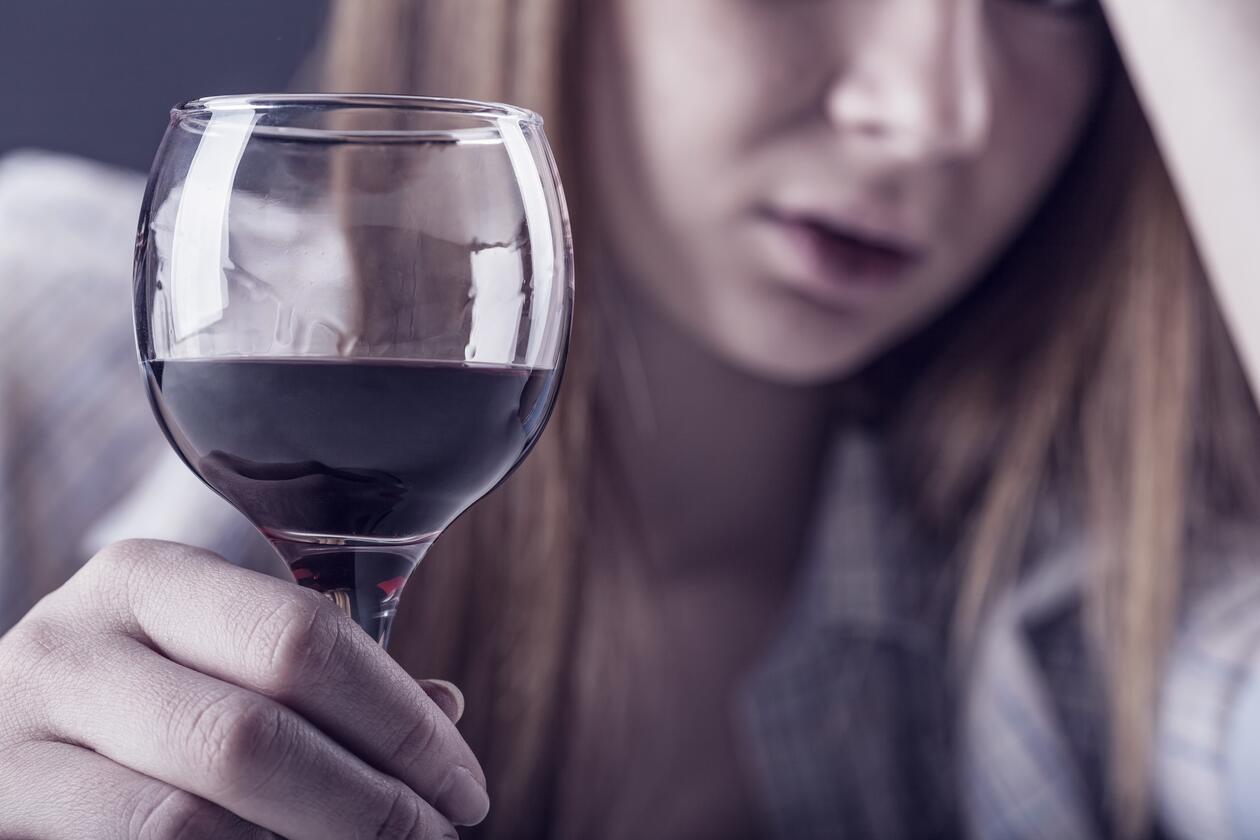Kvinne sitter med hånden i hodet, har et glass rødvin foran seg. Ser oppgitt ut sliten ut