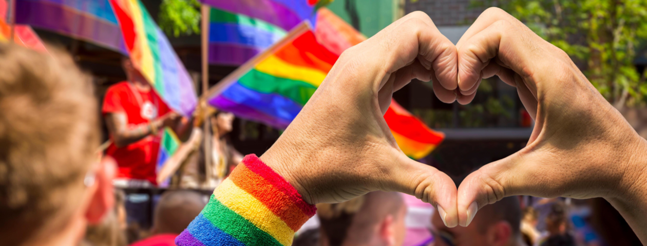 Bilde fra pride parade der noen holder fingrene formet som et hjerte og det er mange regnbueflagg