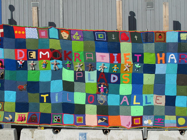 Foto av et heklet lappeteppe ("lappeteppe-grafitti") som gjennom sine ulike motiver på de enkelte rutene skal illustrere demokrati. I tillegg står det skrevet "Demokrati har plads til os alle" med stor skrift i tekstil (dansk).