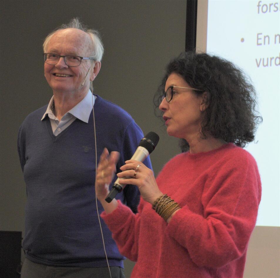 Eldre mann i blå genser står og smiler ved siden av en kvinne i rød genser som snakker i en mikrofon. I bakgrunnen er en skjerm med tekst.