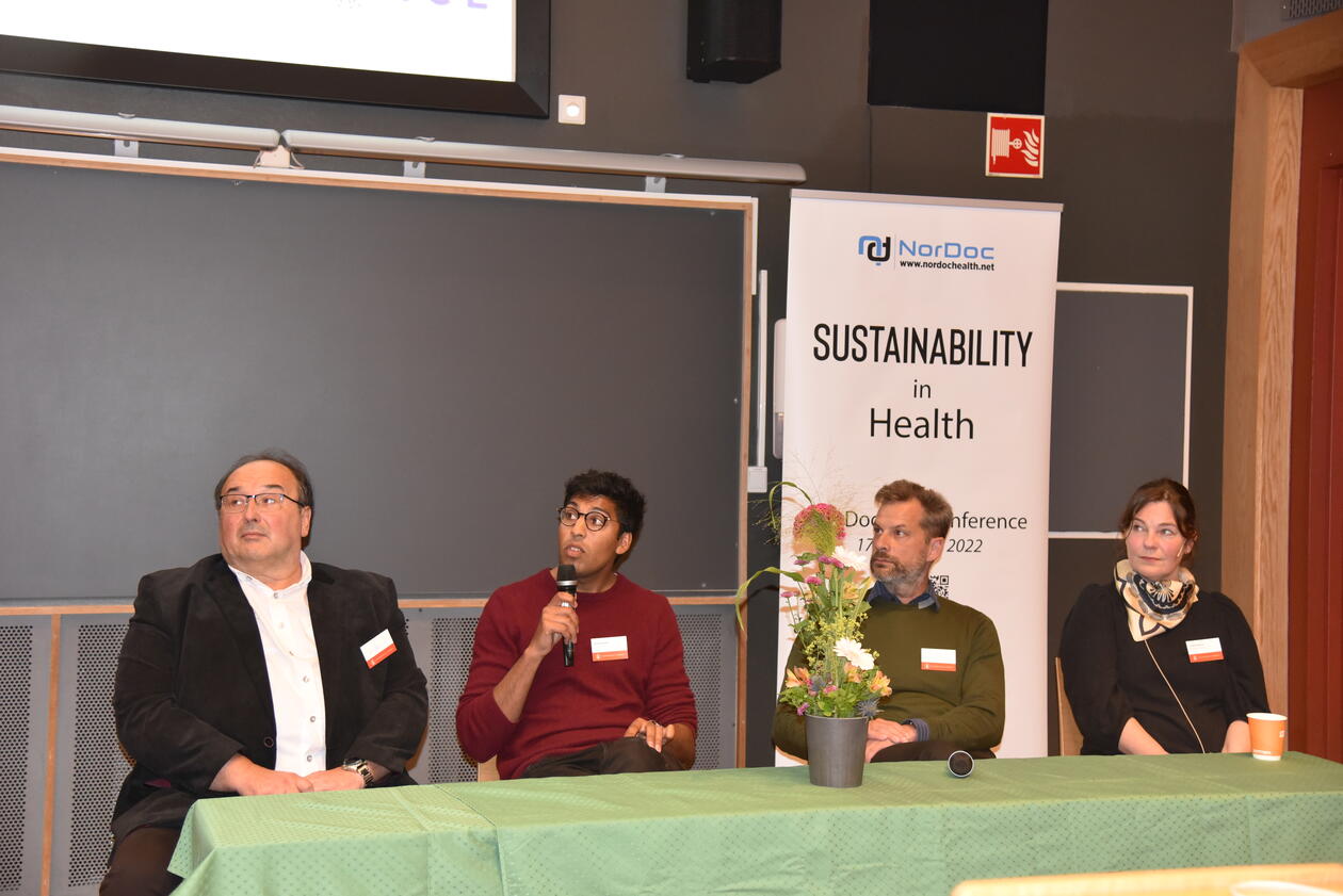 Panel debate Panel debate participants sitting at a green table. PhD. Candidate Anand Bhopal is holding the microfon and saying something
