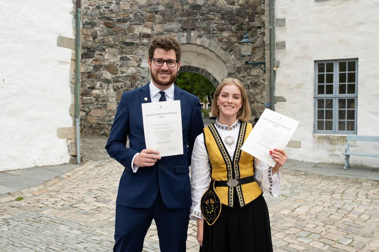 To smilende, festkledde bachelorstudenter, Thomas og Rebekka viser frem sitt diplom. De står ute i borregården ved Håkonshallen.