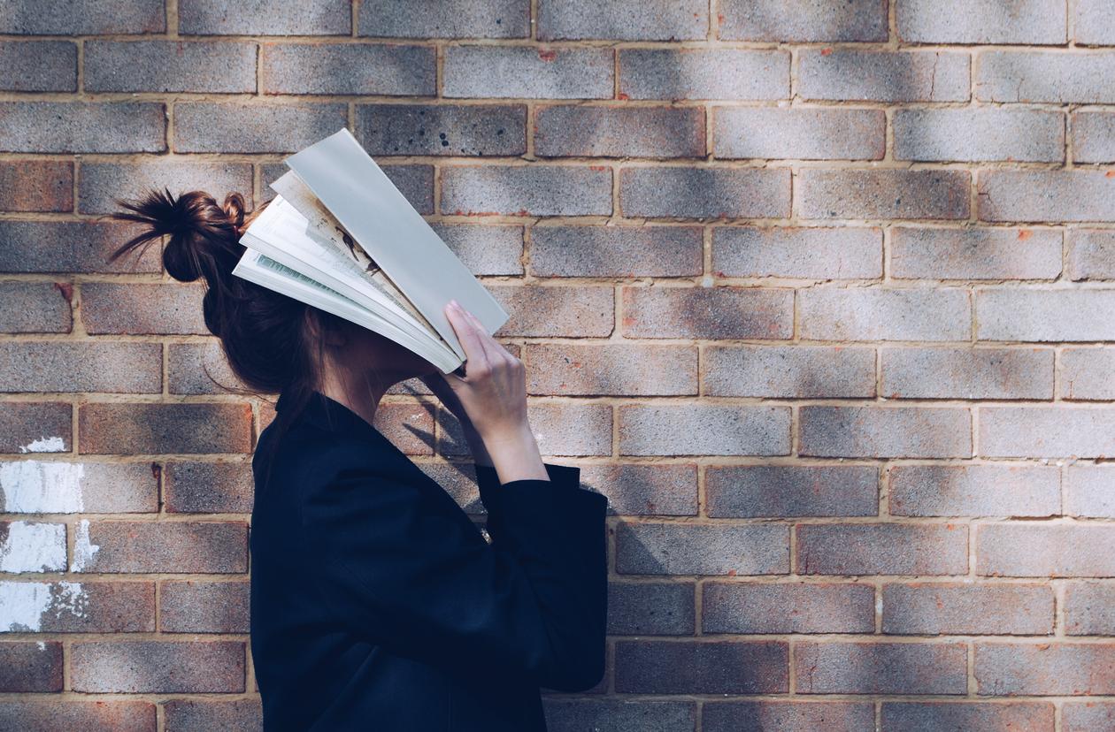 Student in front of natural rustic red brick background holding book up to her face.