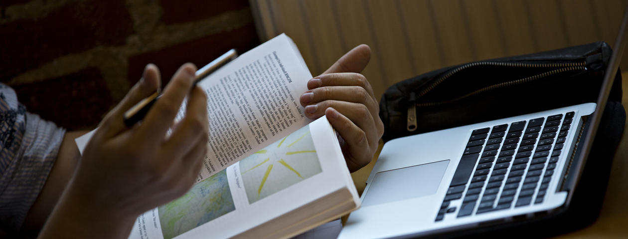 Nærbilde av hender som holder en bok foran en PC
