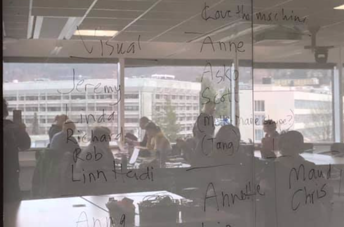 Photo of a wall with names written on it. In the shiny surface of the wall you can see the reflection of people sitting at tables, and a view of buildings seen out of large windows. 