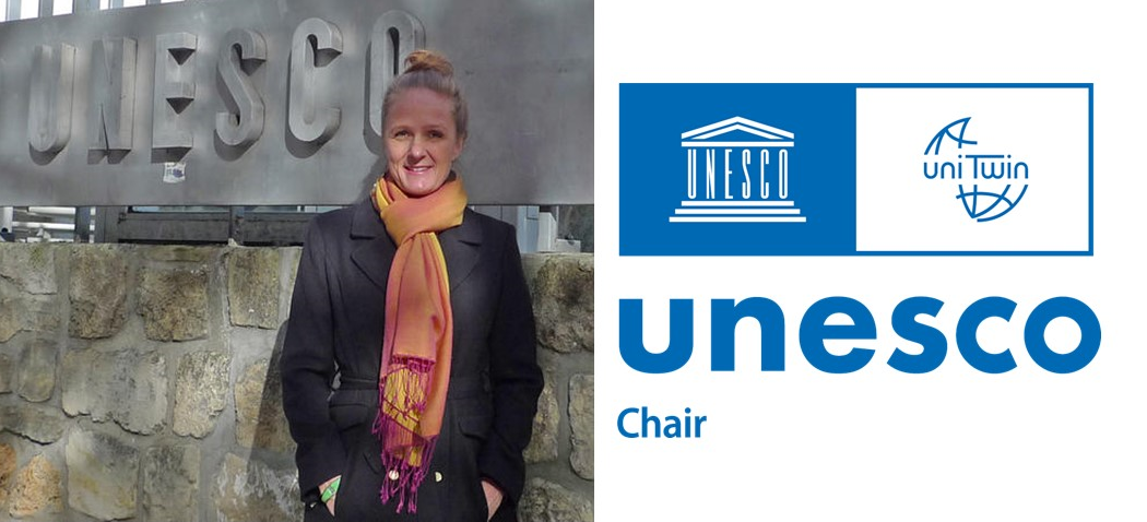 Woman wearing a coat and scarf, smiling in front of large unesco sign. Picture marked with UNESCO chair logo.