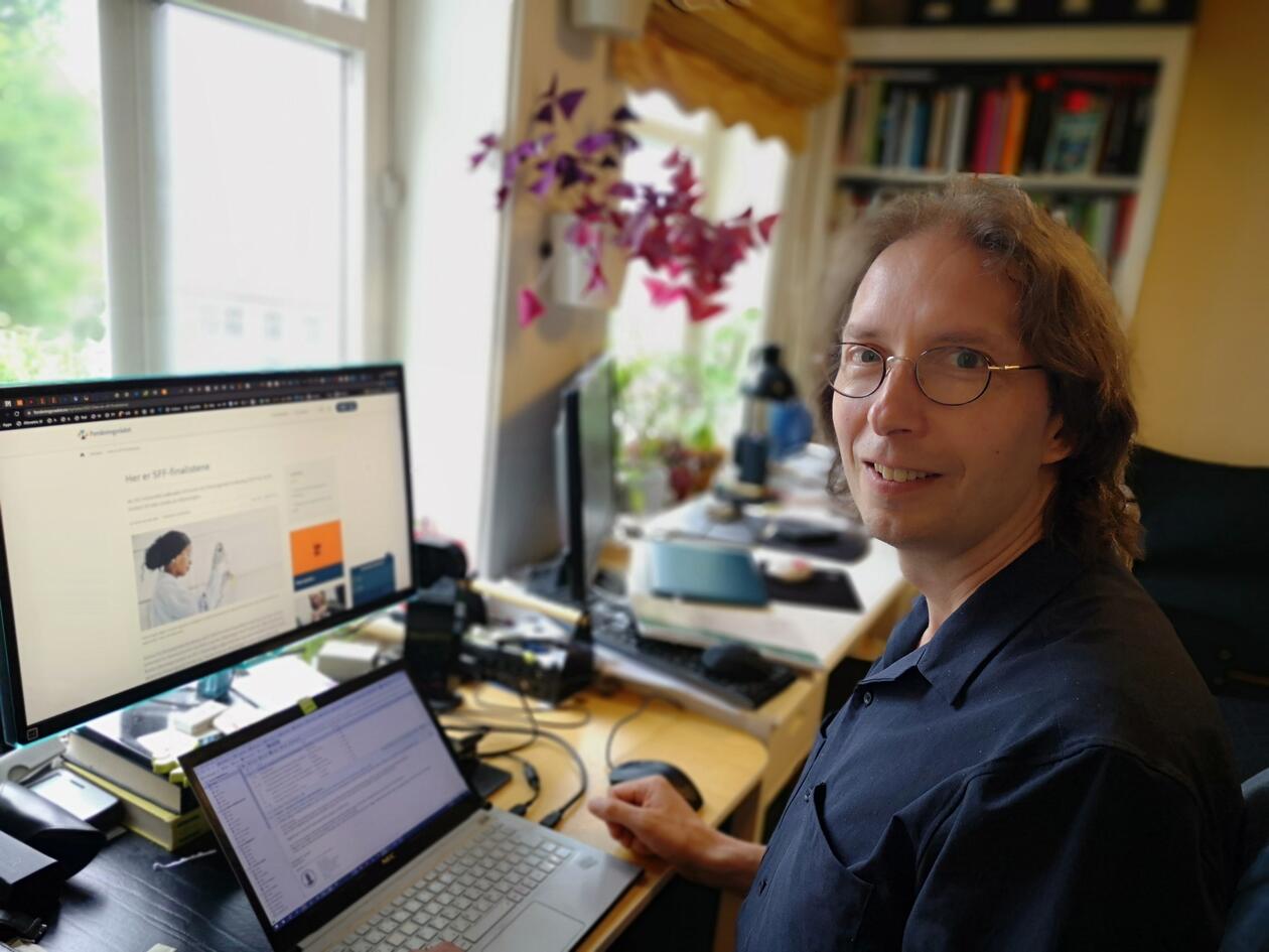 Smilande mann med briler og svart skjorte sit framfor ein laptop og ein ekstern skjerm