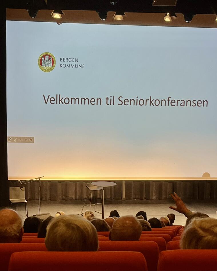 Bildet er tatt i et auditorium. Bildet viser bakhodene på noen av dem som sitter i auditoriet, som ser på et stort lerret fremme på scenen hvor det står "Velkommen til seniorkonferansen"