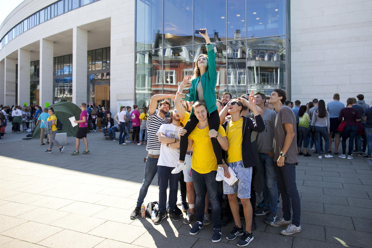 Students in front of the Student Centre standing in a group, carrying one student who's taking a selfie of all of them