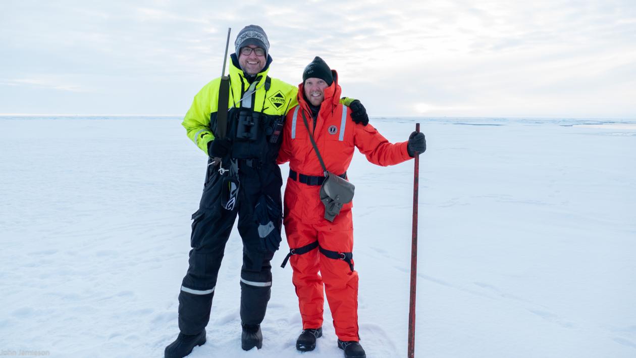 To menn i kjeledress på isen. Alt er hvitt. De smiler til kamera.