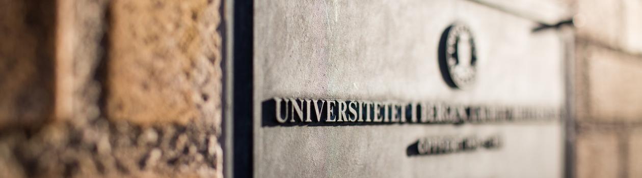 Bilde av en plakett på en murvegg med logoen og navnet til Universitetet i Bergen hvor halve bildet er ute av fokus