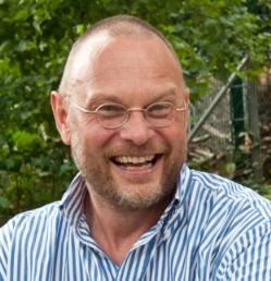 Outdor picture of man with glasses laughing in front of green bushes