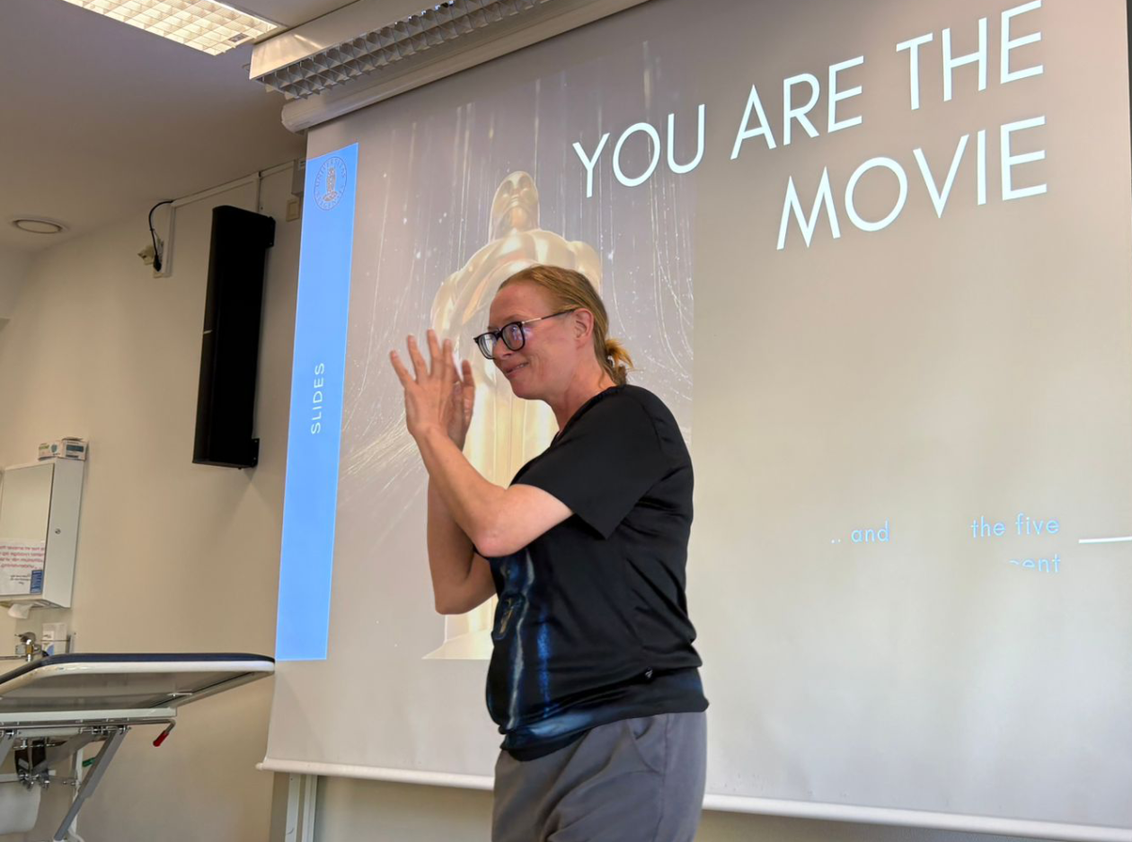 A teacher on the podium waving her hands eagerly. Slide behind her says "You are the movie."
