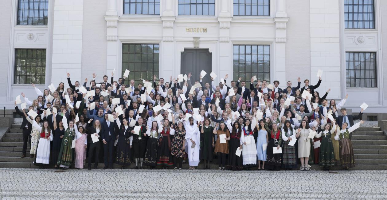 En stor gruppe mennesker i bunad og dress står samlet i trappen forran Naturhistorisk museum. Alle er glade og holder diplomet i hånden i været mens de smiler og ler. Dekanen står fremst i midten av gruppen i dekankappe.