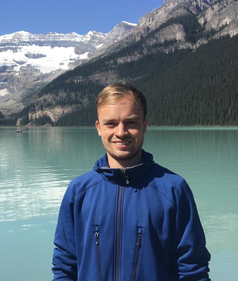 En mann med kort, lyst hår og blå jakke foran en grønn fjord og snøkledde fjell