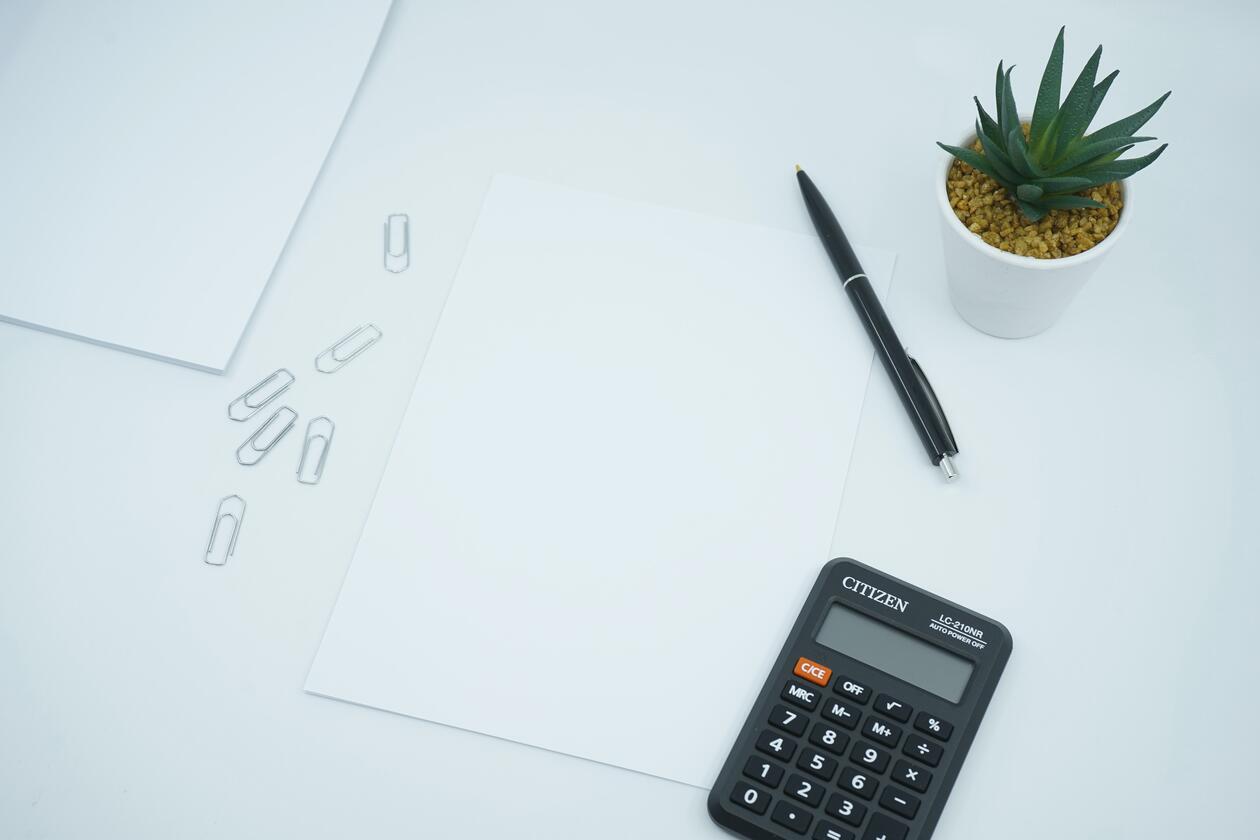 A white table with paperclips, two pieces of white paper, a black pen, a black calculator and a green plant