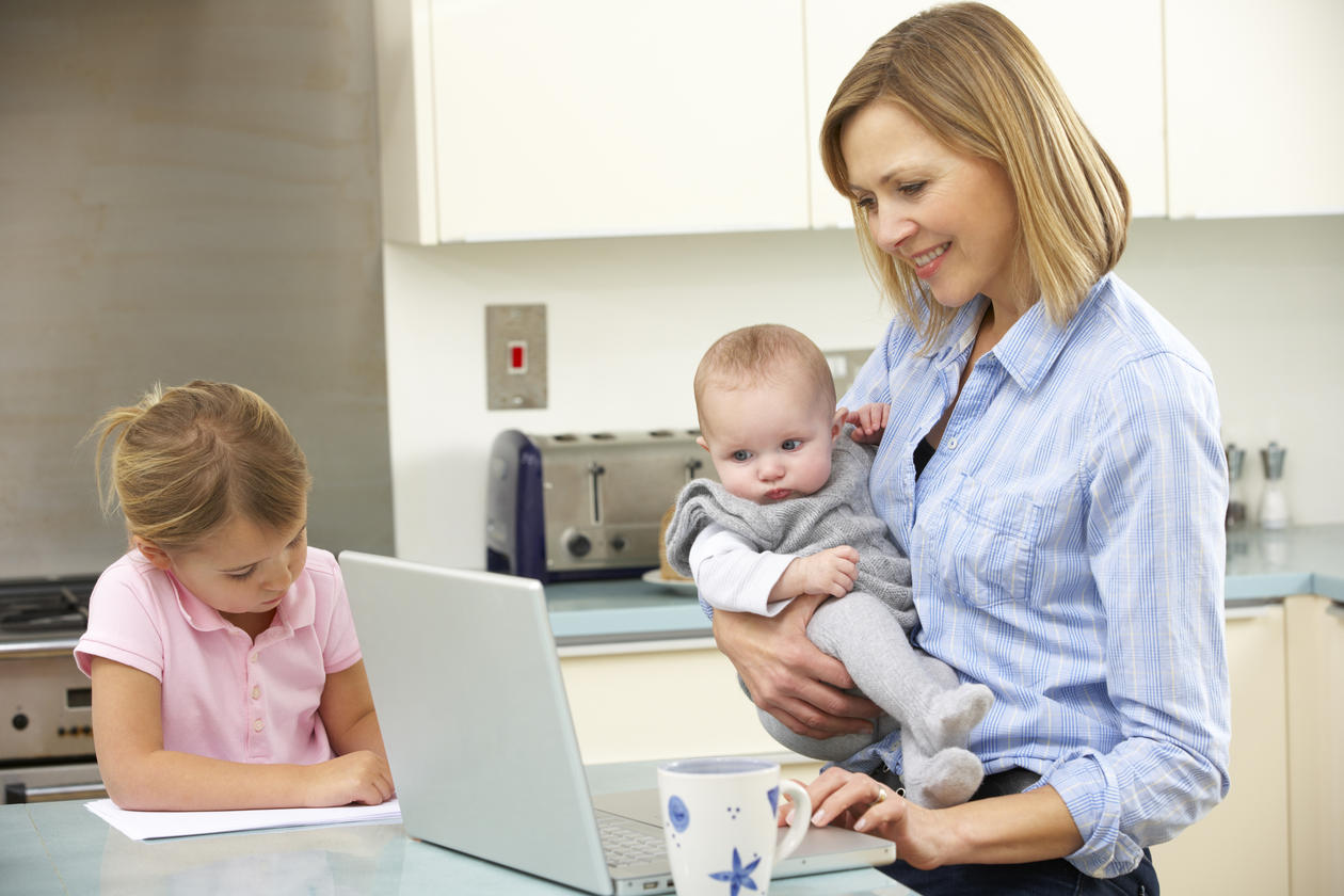 En mor med to barn oppholder seg på kjøkkenet. Mor jobber på datamaskinen mens hun holder minstemann.
