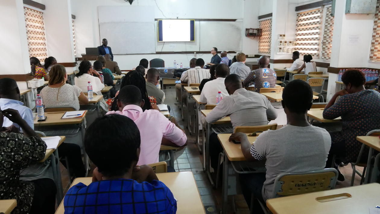 Photo of a classroom in Ghana. We see the backs of many students taking notes, and the professor in the front. 