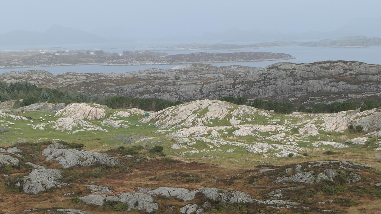 Utsiktsbilde med berg og gressmark i forgrunnen, og sjø og overskyet himmel i bakgrunnnen.