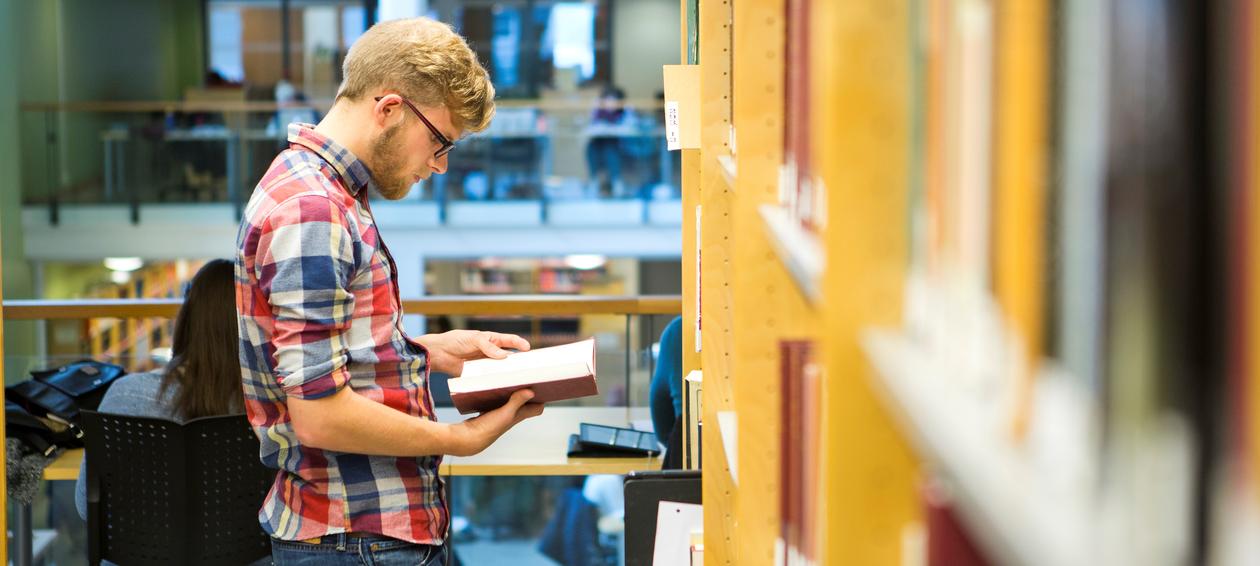 Mannlig student som står og leser ved en bokhylle på biblioteket