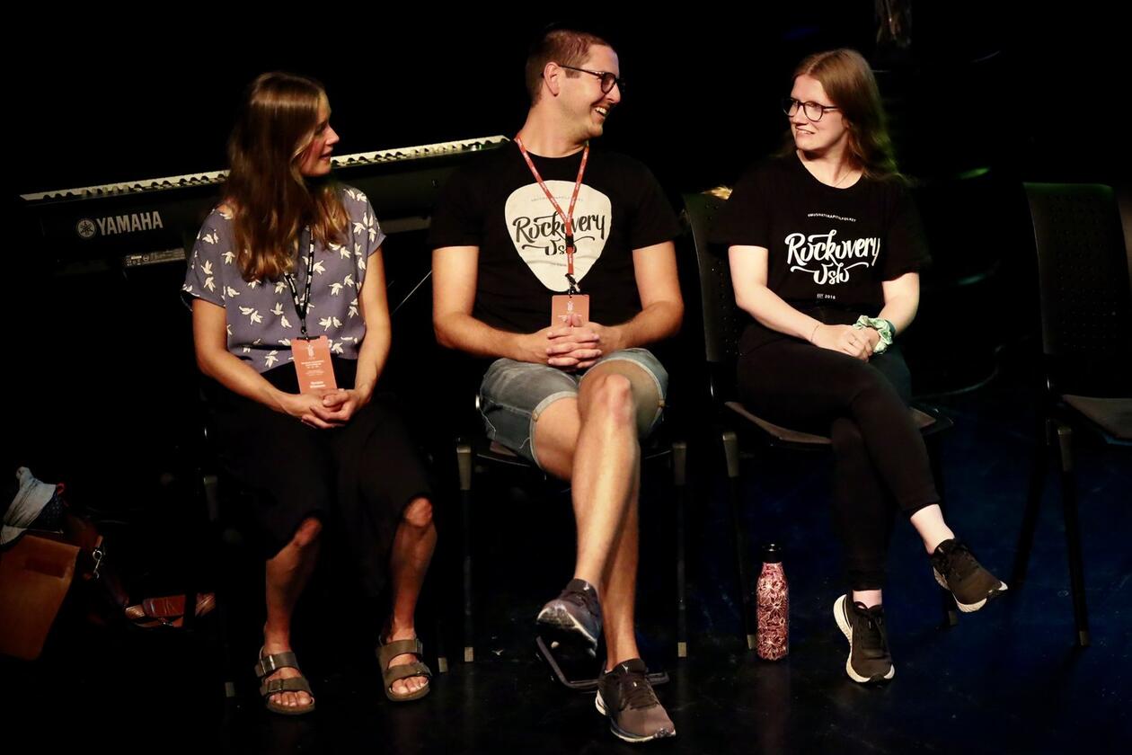 Tre personer sitter ved siden av hverandre på en scene. De ser på hverandre og smiler.