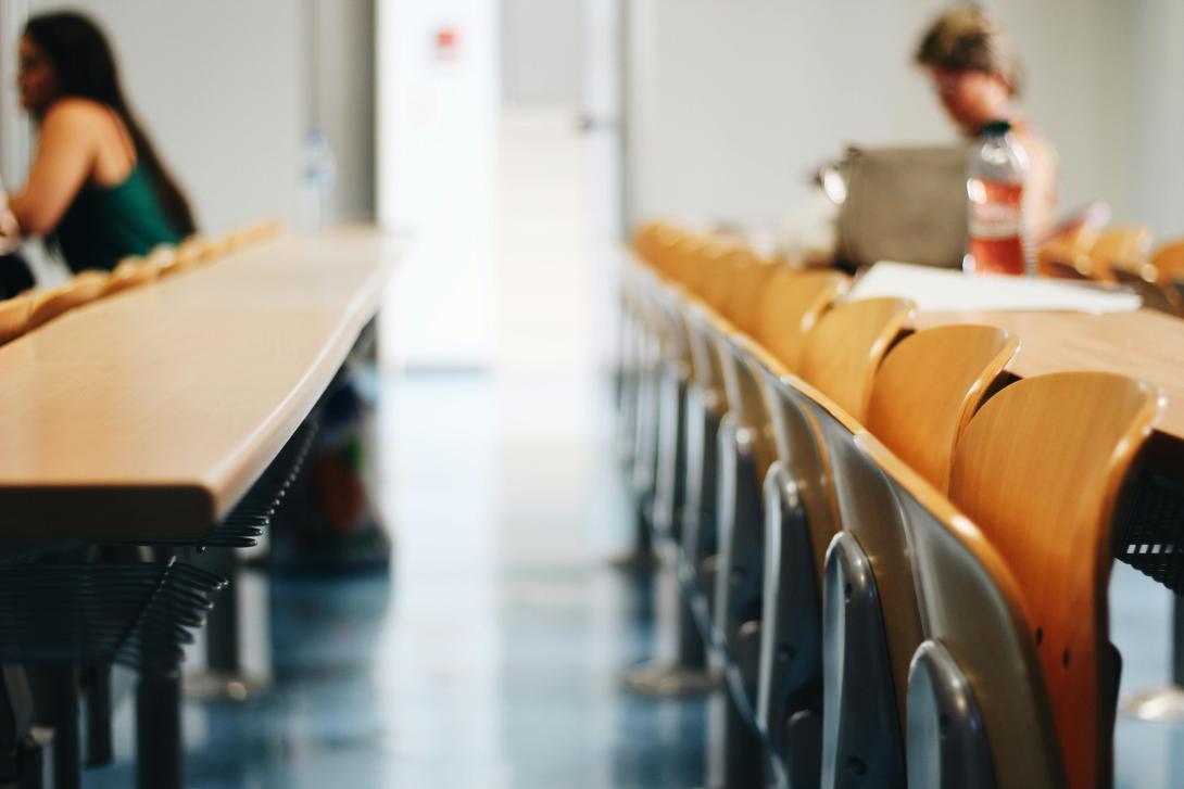 Bildet viser stoler fra et auditorium eller klasserom, raden med stoler er tom og det sitter to personer på raden bak og forran lengst borte fra kamera.