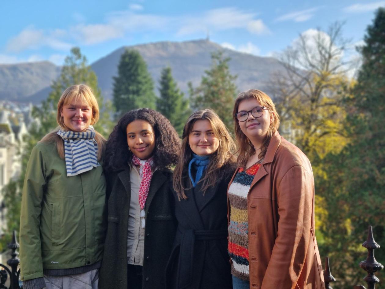 Fire unge kvinner med ytterklede, tre, fjell og blå himmel i bakgrunnen