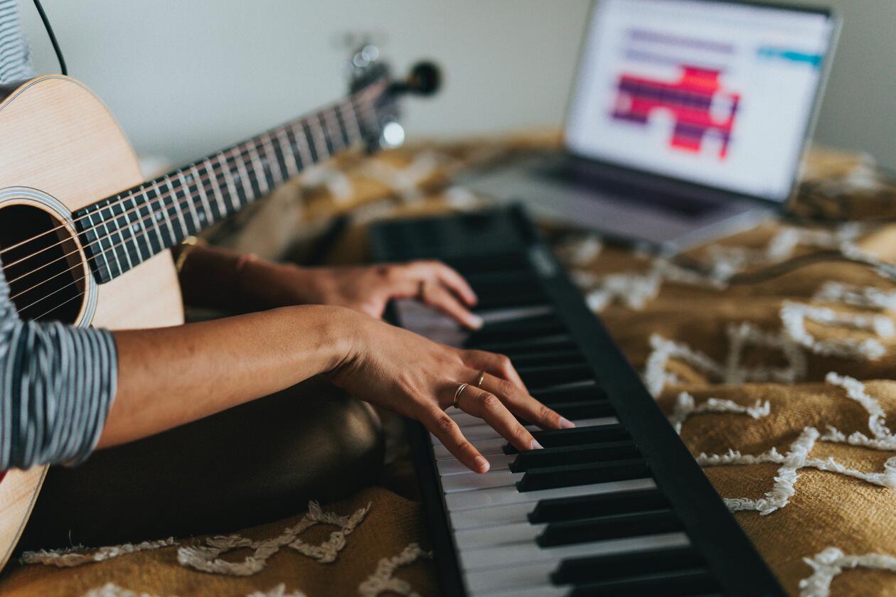 [ID] Bildet viser en person med en guitar i fanget som spiller på et piano. Et brunt teppe er under pianoet og en PC i bakgrunnen.