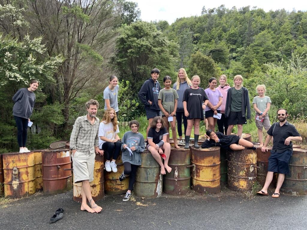 Several people standing and sitting on/by metal barrels on a background of green trees