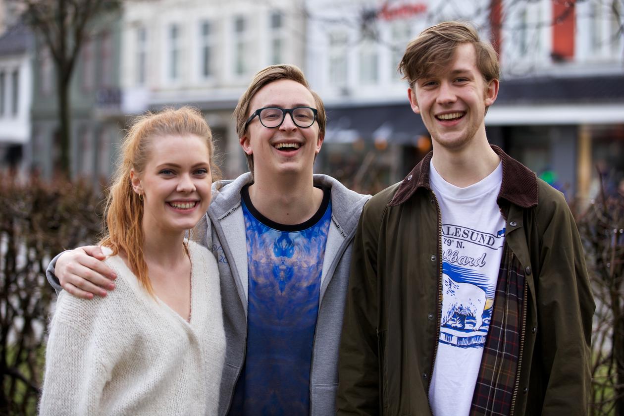 Tre engasjerte studenter smiler mot kamera med hus og park i bakgrunnen.