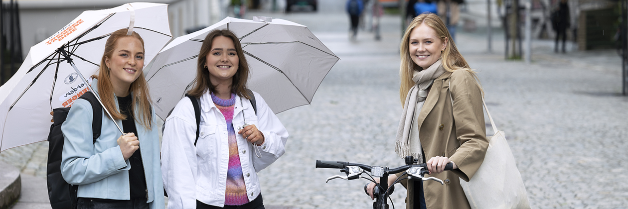 Studenter ute i gaten med paraplyer og sykkel. Tre jenter som smiler mot fotografen