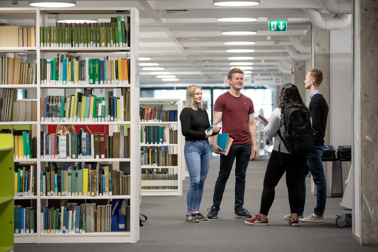 Bilde av 4 studenter som står i biblioteket og snakker sammen