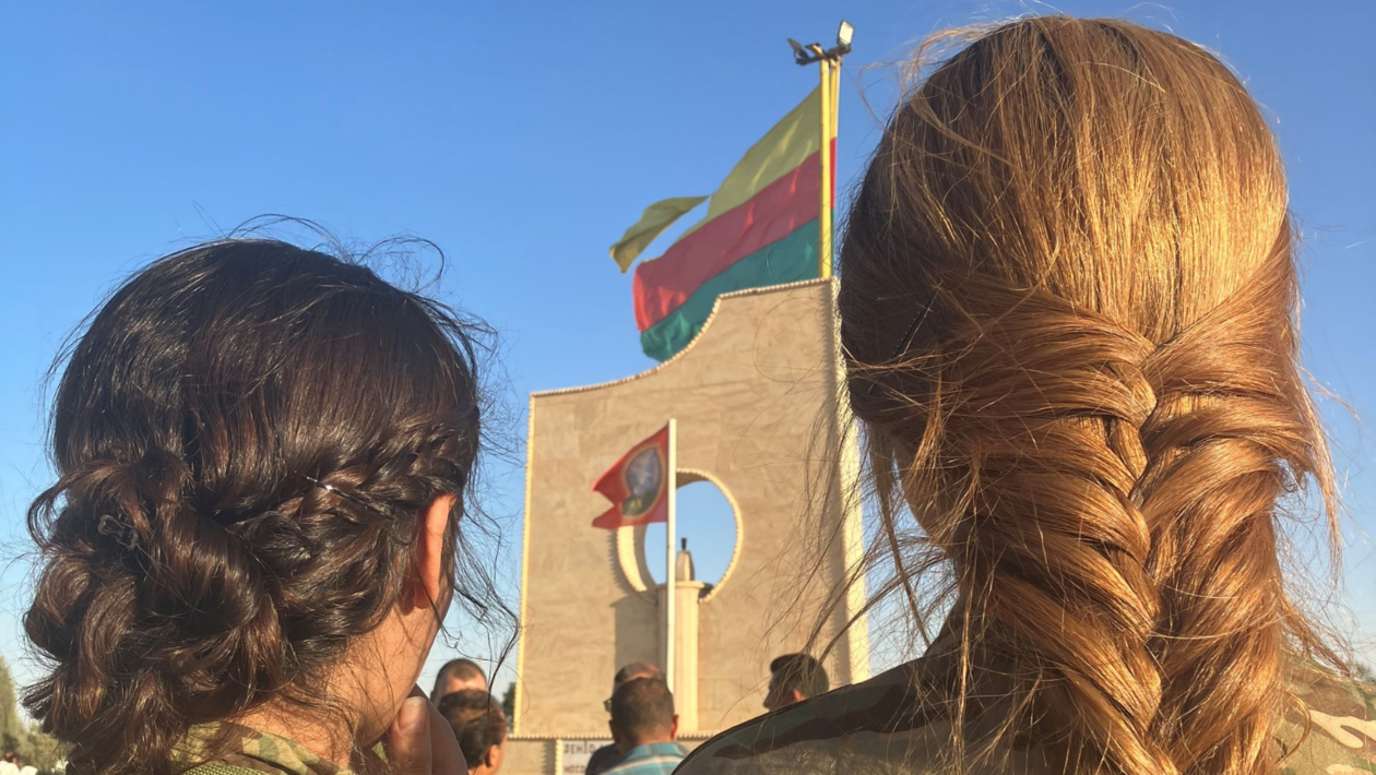 The back head of two women with braided hair, the Rojava flag waving in front of them