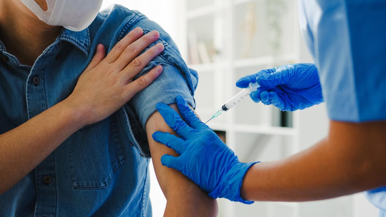 mann i blå trøye og ansiktsmaske får satt en vaksine i armen av en helsearbeider med blå hansker. 