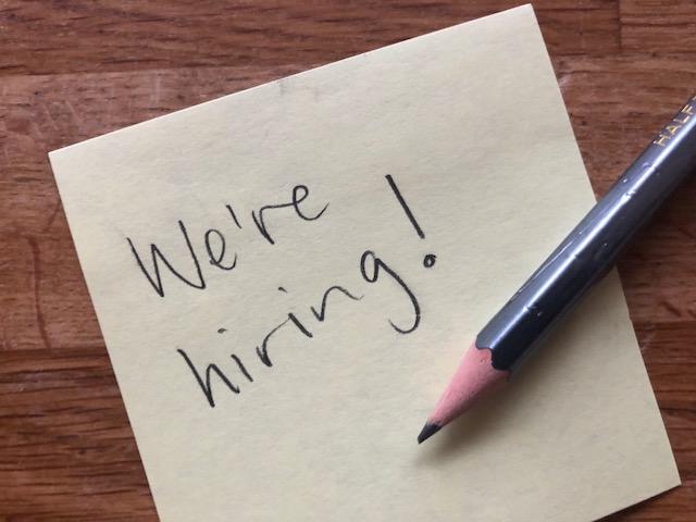 Photo of a post-it note with the handwritten text "we're hiring" written on it.