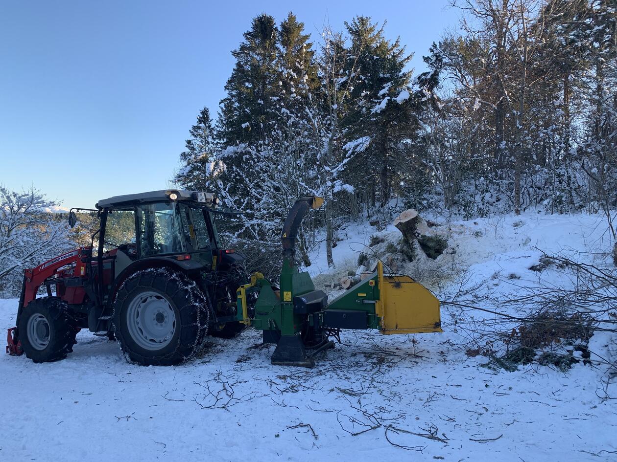 En traktor med vedkutter, tre og blå himmel i bakgrunnen, snø på bakken