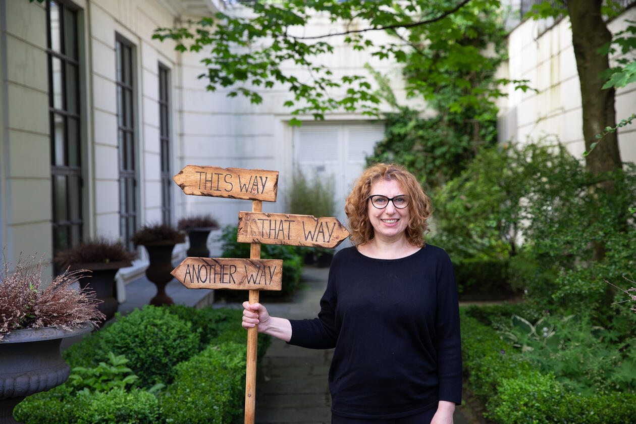 Zhanna Gaulen står i musehagen med et skilt hvor det står "this way" og "that way" som peker i ulike retninger. Frodig grønn bakgrunn, hun har sort genser, rødt krøllete hår og briller og smiler inn i kameraet 