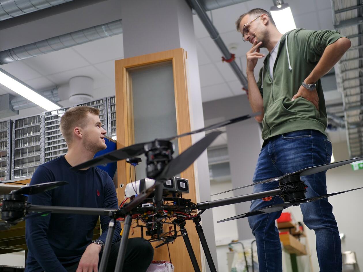 Drone som står på gulvet i et laboratorium. Student og stipendiat snakker i bakgrunnen.
