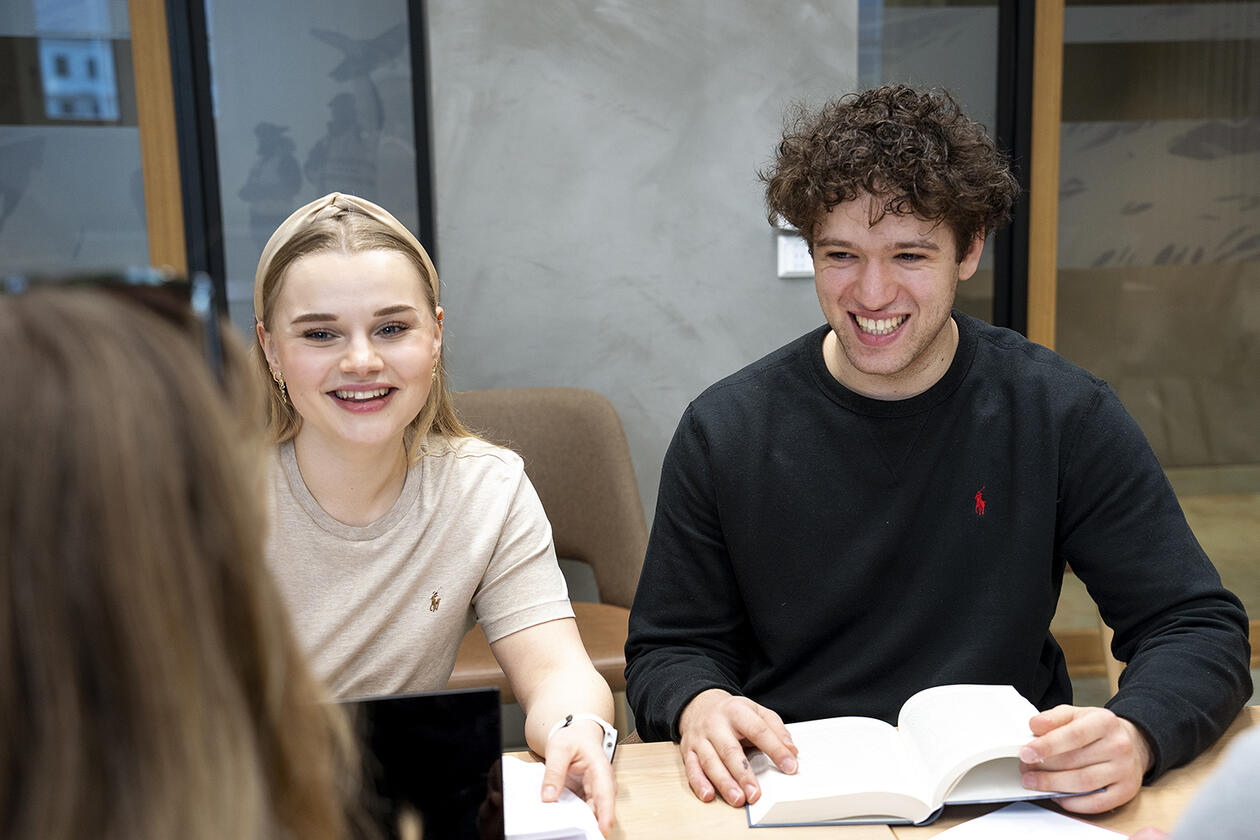To studenter, en jente og en gutt smiler mens de ser på en medstudent. De hviler hendene på en pensumbok og er i en gruppesamarbeidssituasjon