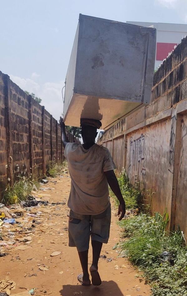 A man balances an old refrigerator on his head, carrying it through a narrow alley leading into Ruga