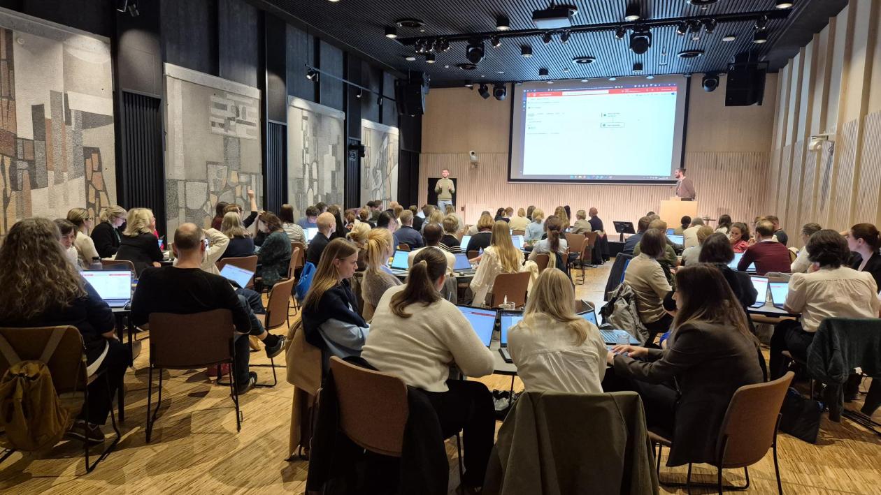 Ansatte som sitter i grupper og jobber i Storsalen, person på scenen presenterer Power Automate